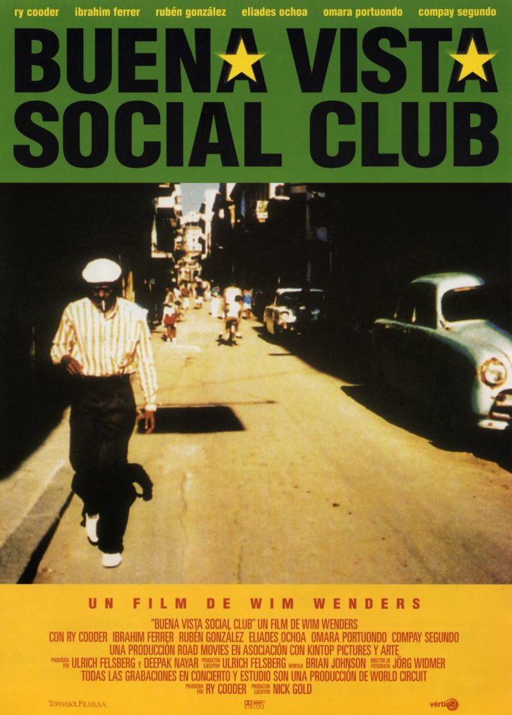 An older gentleman in a striped shirt, kangol cap, and black slacks walks towards us. He has his head lowered and he smokes a cigarette. There's a bustling street behind him and an old Studebaker across from him.